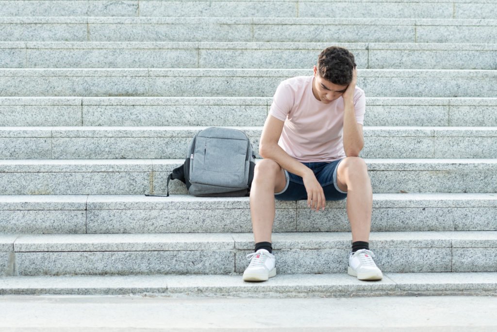 Sad and frustrated hispanic teenager sitting on stairs and holding his head. Anxiety and depression in adolescence concept.